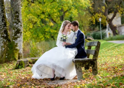 Brautpaar sitzt auf Bank im Herbstlaub, Hochzeit in Bernried festgehalten, liebevoller Moment und Hochzeitsfotografie.