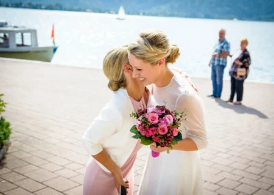 Braut umarmt Gast mit Blumenstrauss bei Hochzeit am Tegernsee. Hochzeitfotografie vor malerischer Seelandschaft.