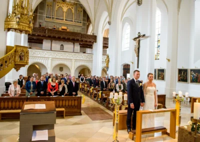 Brautpaar bei kirchlicher Trauung in Stadtpfarrkirche Maria Himmelfahrt, umgeben von Gästen. Hochzeitsfoto in Bayern.