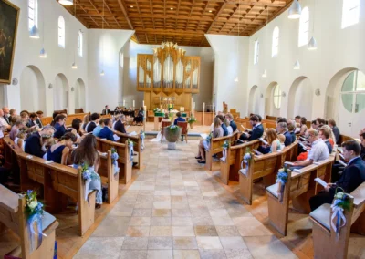 Hochzeitsfeier in Kirche in München; Gäste sitzen in Bänken, dekoriert mit Blumen und Bändern. Hochzeitsreportage.