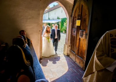 Braut in weissem Kleid mit Brautvater betritt Kirche zur Trauung in Obermenzing, festgehalten bei einer Hochzeitsreportage.