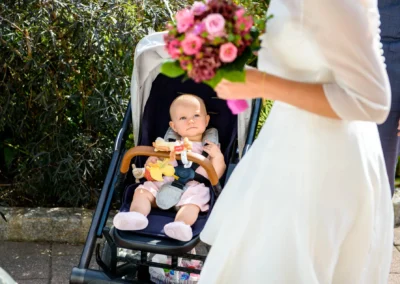 Baby im Kinderwagen schaut auf Braut mit Blumenstrauss bei Hochzeit im Rathaus Tegernsee. Hochzeitsreportage in Bayern.