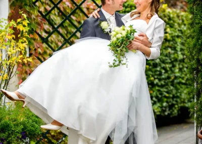 Brautpaar in Pullach, der Bräutigam hebt die Braut vor einem Laubtor auf. Hochzeitsfotografie in Pullach im Isartal.