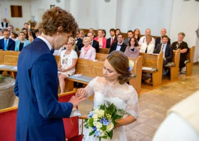 Brautpaar beim Ringtausch in Kirche in Sendling, München. Hochzeitsreportage mit Blumenstrauss und Gaesten.