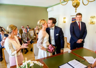 Brautpaar beim Kuss im Rathaus Tegernsee, umgeben von applaudierenden Hochzeitsgaesten. Hochzeitsreportage Bayern.