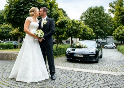 Brautpaar bei der Hochzeit in St. Michael, Perlach, vor einem dekorierten Auto auf Kopfsteinpflaster.