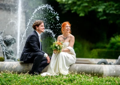Brautpaar bei Hochzeitsportraits am Friedensengel in München, lacht vor einem Springbrunnen im Gruenen. Hochzeitsfotografie.