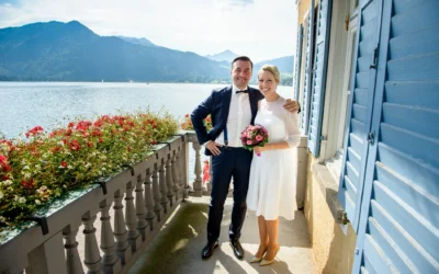 Brautpaar laechelt auf Balkon mit Blick auf Tegernsee, umgeben von Blumen. Hochzeitfotografie Tegernsee.