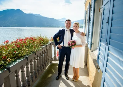 Brautpaar laechelt auf Balkon mit Blick auf Tegernsee, umgeben von Blumen. Hochzeitfotografie Tegernsee.