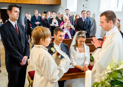 Kirchliche Trauung in Solln: Brautpaar kniet, Priester segnet, Gäste schauen zu. Hochzeitsfotografie in München.