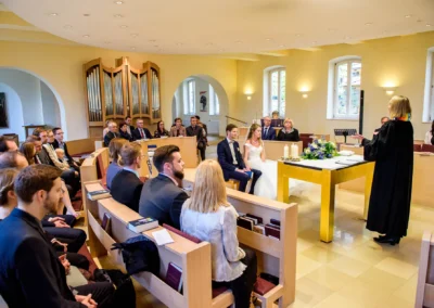 Kirchliche Trauung in Bernried: Brautpaar vor der Traurednerin, Gaeste sitzen in einer Kirche. Hochzeitsfotografie Bayern.
