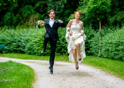 Brautpaar springt fröhlich auf Weg bei Hochzeitsportraits im Grünen, München, festgehalten von Hochzeitsfotograf.