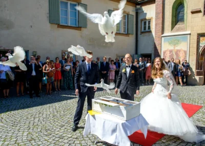 Brautpaar laesst weisse Tauben fliegen, Hochzeitsfoto Schloss Blutenburg Oberbayern, Gaeste im Hintergrund.