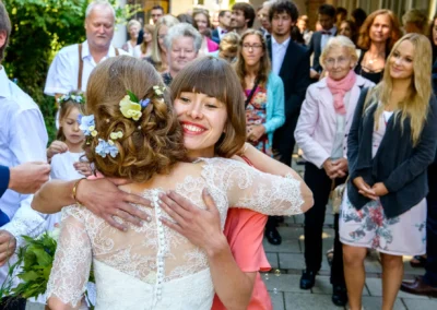 Braut umarmt Gast bei Hochzeit in München, Hochzeitsreportage, freudiger Moment mit Blumen und Gaesten im Hintergrund.