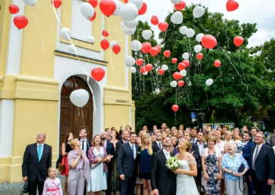 Hochzeit in St. Michael, Perlach: Brautpaar und Gaeste lassen rote und weisse Luftballons steigen. Hochzeitsfotografie.