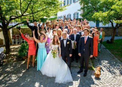 Gruppenfoto bei einer Hochzeit in Oberbayern. Brautpaar mit Gaesten vor Schloss Blutenburg in fröhlicher Stimmung.
