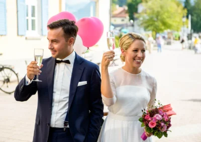 Bräutigam und Braut mit Sekt bei Hochzeit vor dem Rathaus Tegernsee. Hochzeitsfotografie mit Blumenstrauss und Luftballons.