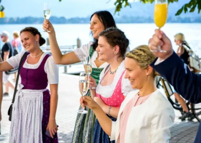 Gäste in Dirndl stoßen bei einer Hochzeit am Tegernsee an, im Hintergrund der See. Hochzeitsfotografie in Bayern.