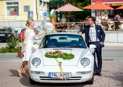 Brautpaar posiert an dekoriertem Auto vor Rathaus Tegernsee. Perfekte Hochzeitsfotografie in Bayern.
