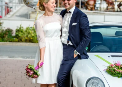 Brautpaar steht lachend neben Auto mit Blumen, bei Hochzeit in Rottach-Egern, inklusive Just Married Ballon.