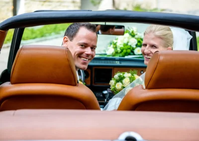 Brautpaar lacht im Cabrio mit Blumenstrauss, Brautpaarfotos im Tegernsee Hotel, Hochzeitsreportage Oberbayern.