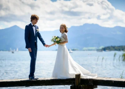 Brautpaar am Chiemsee-Steeg, umgeben von Bergen und Wasser. Hochzeitsreportage Oberbayern für Hochzeitsfotograf.