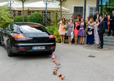 Hochzeitsreportage: Gruppe winkt neben Auto mit Blechdosen an der Hochzeit nahe Muenchen. Festliche Stimmung.