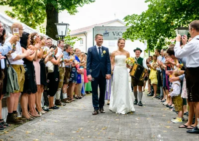 Brautpaar bei Hochzeitsfeier in ViCulinaris, mit Gaesten im traditionellen bayerischen Outfit und Akkordeonspieler.
