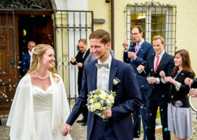 Brautpaar laechelnd vor Kirche, umgeben von Gaesten mit Seifenblasen, Hochzeit Bernried, festgehalten vom Hochzeitsfotografen.