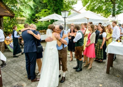 Hochzeitsfeier in Bad Toelz: Braut in weissem Kleid umarmt Gaeste. Fröhliche Gesellschaft feiert im Freien.