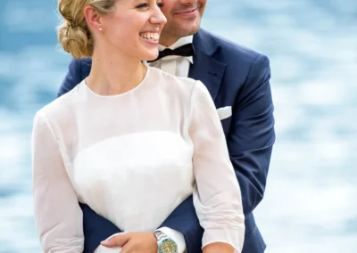 Brautpaar laechelnd vor dem Tegernsee bei der Hochzeit. Der Bräutigam umarmt die Braut, die einen Blumenstrauss haelt.