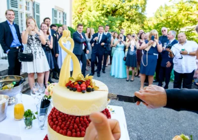 Hochzeitsfeier in Oberbayern: Brautpaar schneidet Torte an, Gaeste schauen gespannt bei der Trauung zu.