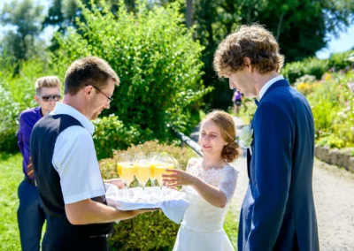 Hochzeit am Chiemsee: Brautpaar bei der Feier im Freien, Sektempfang mit Sonnenlicht und gruenem Garten.