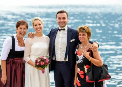 Glückliches Brautpaar mit Gästen am Tegernsee. Hochzeitsfotografie zeigt strahlende Gesichter vor malerischer Seekulisse.
