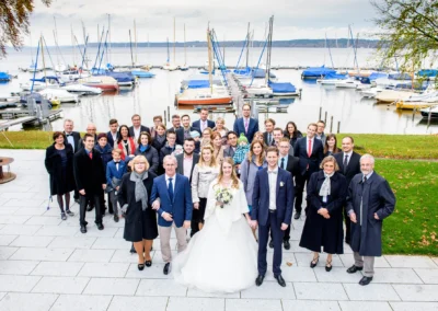 Hochzeitsreportage in Bernried: Brautpaar mit Gaesten am Starnberger See, festlich gekleidete Gruppe mit Segelbooten im Hintergrund.