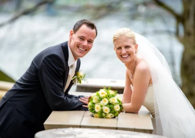 Brautpaar lachend mit Blumenstrauss am Tegernsee. Hochzeitsreportage im Tegernsee Hotel, suedlich von Muenchen.
