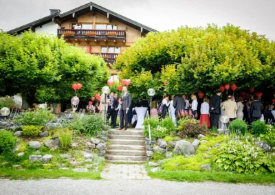 Hochzeitsfeier in Bad Wiessee mit Gästen und roter Ballondeko vor einem traditionellen Gebäude, umgeben von Bäumen.