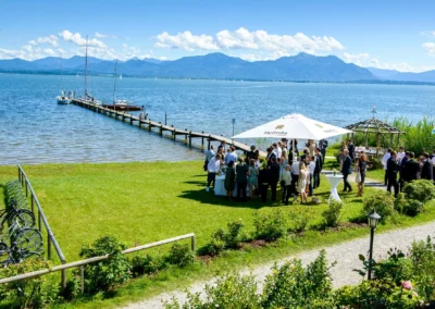 Hochzeitsfeier am Chiemsee mit Gaesten unter Sonnenschirmen, Bergkulisse im Hintergrund, Trauung im Freien.