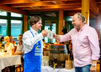 Zwei Maenner stoßen bei der Hochzeit im Wirtshaus am Rosengarten in Muenchen mit Bierkruegen an, Gaeste im Hintergrund.