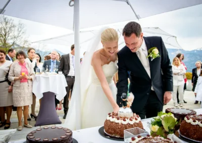 Brautpaar schneidet Hochzeitstorte bei Feier im Tegernsee Hotel, umgeben von Gaesten und Bergen in Oberbayern.