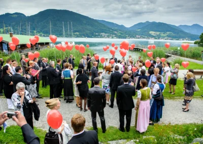 Hochzeitsgesellschaft mit roten Luftballons am Tegernsee bei Feier im Freien, Berge im Hintergrund. Hochzeitsreportage.
