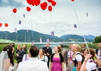 Hochzeitsgaeste lassen rote Ballons am Tegernsee steigen, festliche Trauung im Freien, Hochzeitsreportage.