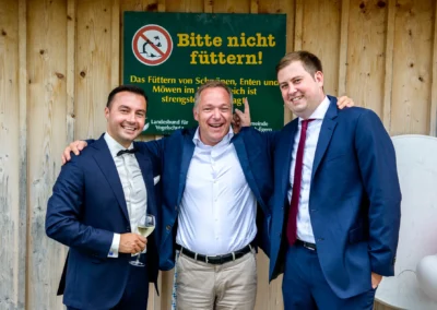 Drei lachende Maenner in Anzuegen bei Hochzeit in Rottach-Egern vor Holzverkleidung und Schild. Hochzeitsreportage.