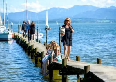 Hochzeitsgesellschaft am Steg am Chiemsee, Kinder sitzen am Ufer, Segelboote im Hintergrund, Oberbayern.