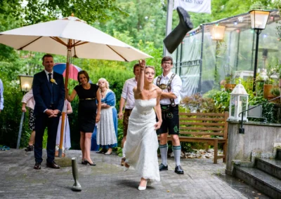 Hochzeitsfoto bei ViCulinaris: Braut wirft Gummistiefel bei Hochzeitsfeier in Bad Toelz, umgeben von festlichen Gaesten.