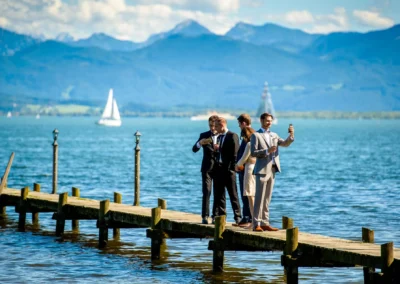 Hochzeitsgaeste machen Fotos am Steg vor Bergkulisse am Chiemsee, Hochzeitsreportage in Oberbayern.