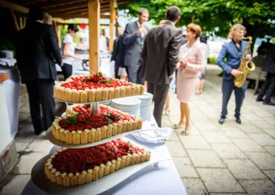 Hochzeitsfeier in Bad Wiessee mit mehrstoeckiger Erdbeertorte, Gaeste im Hintergrund und Saxophonspieler.