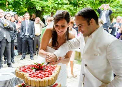Brautpaar schneidet Hochzeitstorte bei Hochzeitsfeier in Bad Wiessee, umgeben von Gaesten im Terrassenhof.