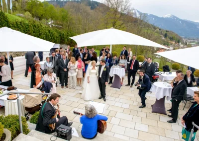 Gartenhochzeit im Tegernsee Hotel, Gaeste lachen, Musiker spielen Gitarre, Brautpaar im Mittelpunkt, Alpenhintergrund.
