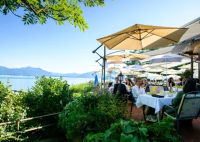 Hochzeitsfeier am Chiemsee mit Blick auf Berge. Gaeste sitzen unter Sonnenschirmen beim Empfang. Hochzeitsreportage.